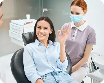 Smiling patient making an “OK” gesture while seated in a dental chair, with a masked dental professional nearby—capturing confidence, comfort, and a successful dental visit